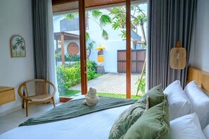 A bedroom with a bed, a chair, a small statue, and a view of a garden and building through a glass door. - Villa Moon Orchid - 3 bedroom villa (Bali)