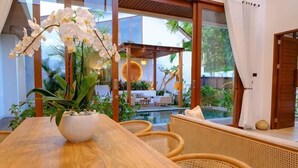 A wooden dining table with a white vase of orchids, wicker chairs, and a view of a pool and outdoor seating area.