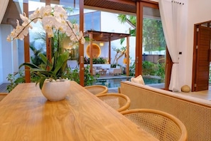 A wooden dining table with a white vase of orchids, wicker chairs, and a view of a pool and outdoor seating area. - Villa Moon Orchid - 3 bedroom villa (Bali)