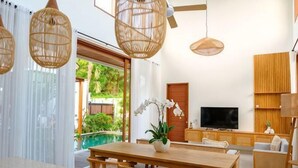 A modern living room with a wooden dining table, wicker chairs, a TV, and a large window with a view of greenery.