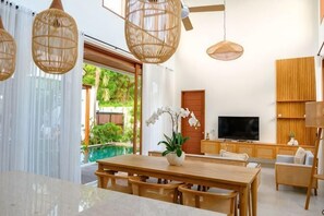 Una sala de estar moderna con una mesa de comedor de madera, sillas de mimbre, un televisor y un ventanal con vistas a zonas verdes.