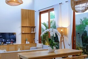 A modern living room with a wooden dining table, wicker furniture, a television, and a large window with curtains. - Villa Moon Orchid - 3 bedroom villa (Bali)