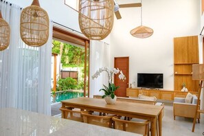 Una sala de estar moderna con una mesa de comedor de madera, sillas de mimbre, un televisor y un amplio ventanal con vistas a la vegetación.