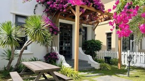 Une terrasse avec une table en bois et des bancs, entourée d’une végétation luxuriante et de plantes en fleurs.