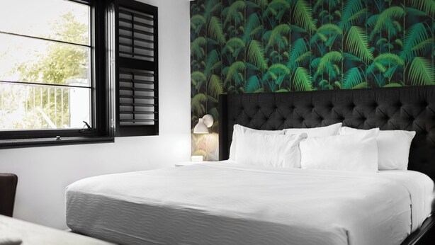 Una cama bien tendida con sábanas blancas en una habitación con un ventanal grande y una pared decorada con un diseño de hojas tropicales.