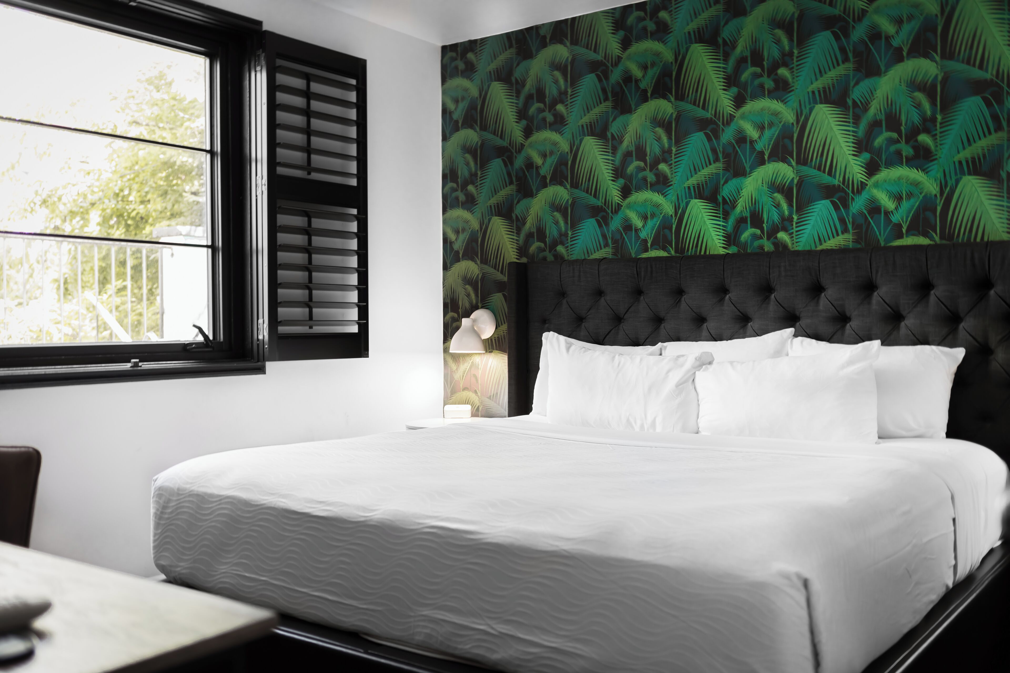 A neatly made bed with white linens in a room with a large window and a wall with a tropical leaf pattern.