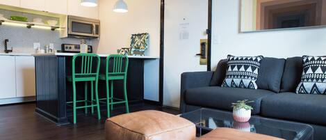 A modern kitchen with a microwave, green chairs, and a dining area. There is a sofa with patterned cushions and a coffee table with a plant.