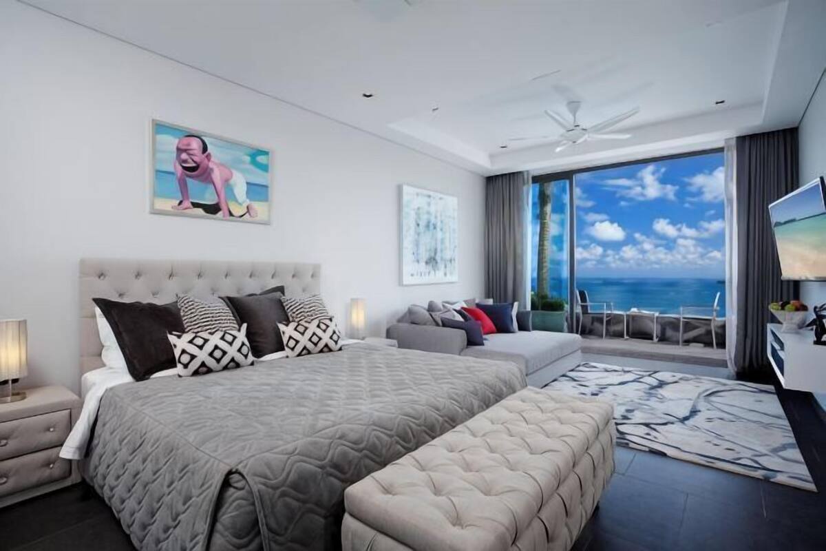 A modern bedroom with a large bed, a sofa, a TV, and a balcony with a view of the sea.