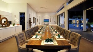 A modern dining room with a long table set for a meal, a kitchen area with white cabinets, and a view of the outdoors.