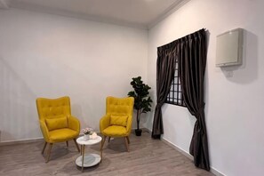 Een kamer met twee gele stoelen, een klein rond tafeltje met een vaas, een potplant en een raam met gordijnen.