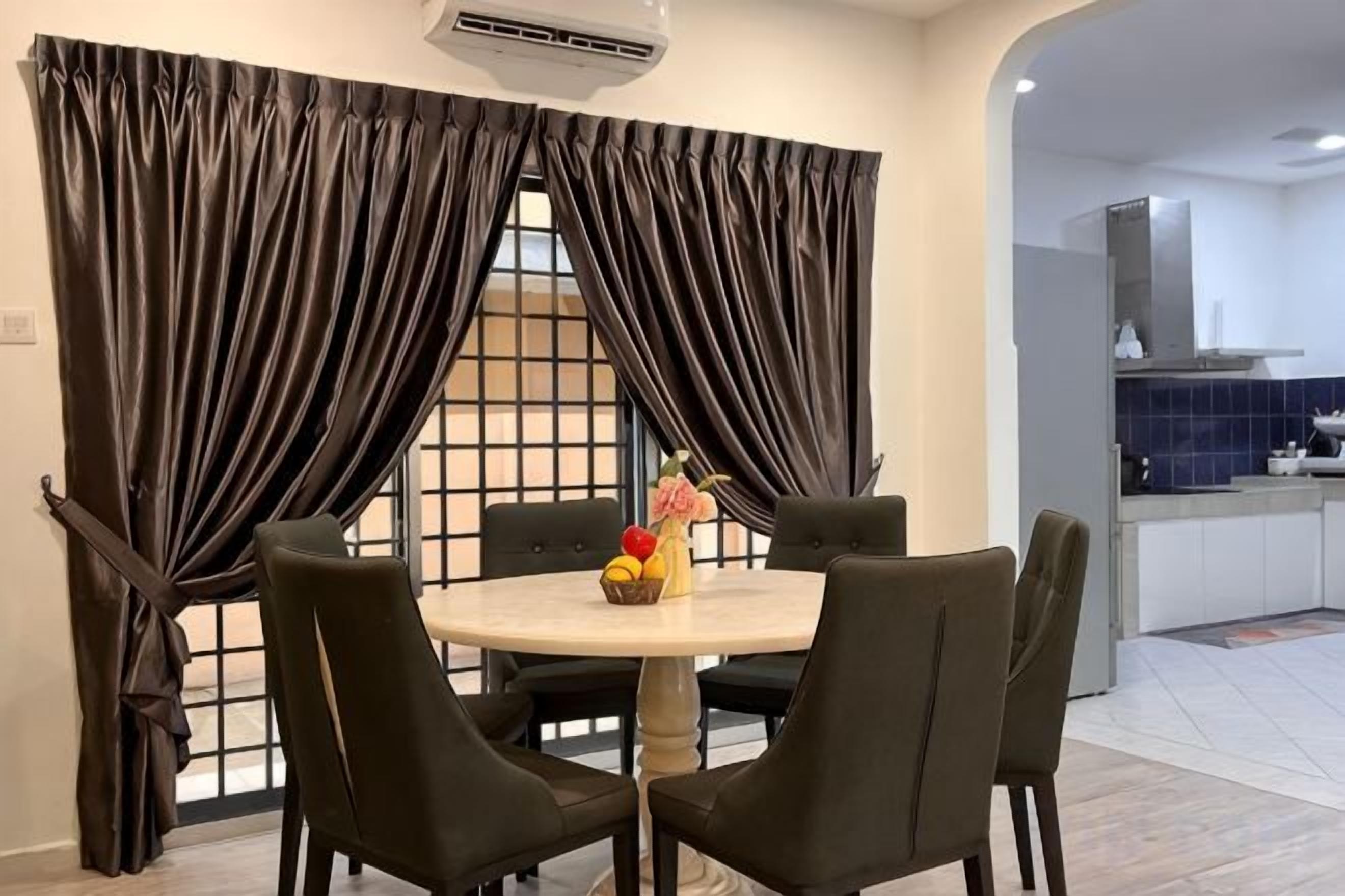 A dining area with a round table and four chairs, a window with curtains, and a kitchen in the background.