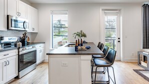 A modern kitchen with white cabinets, stainless steel appliances, a central island with bar stools, and large windows offering views of the outdoors.