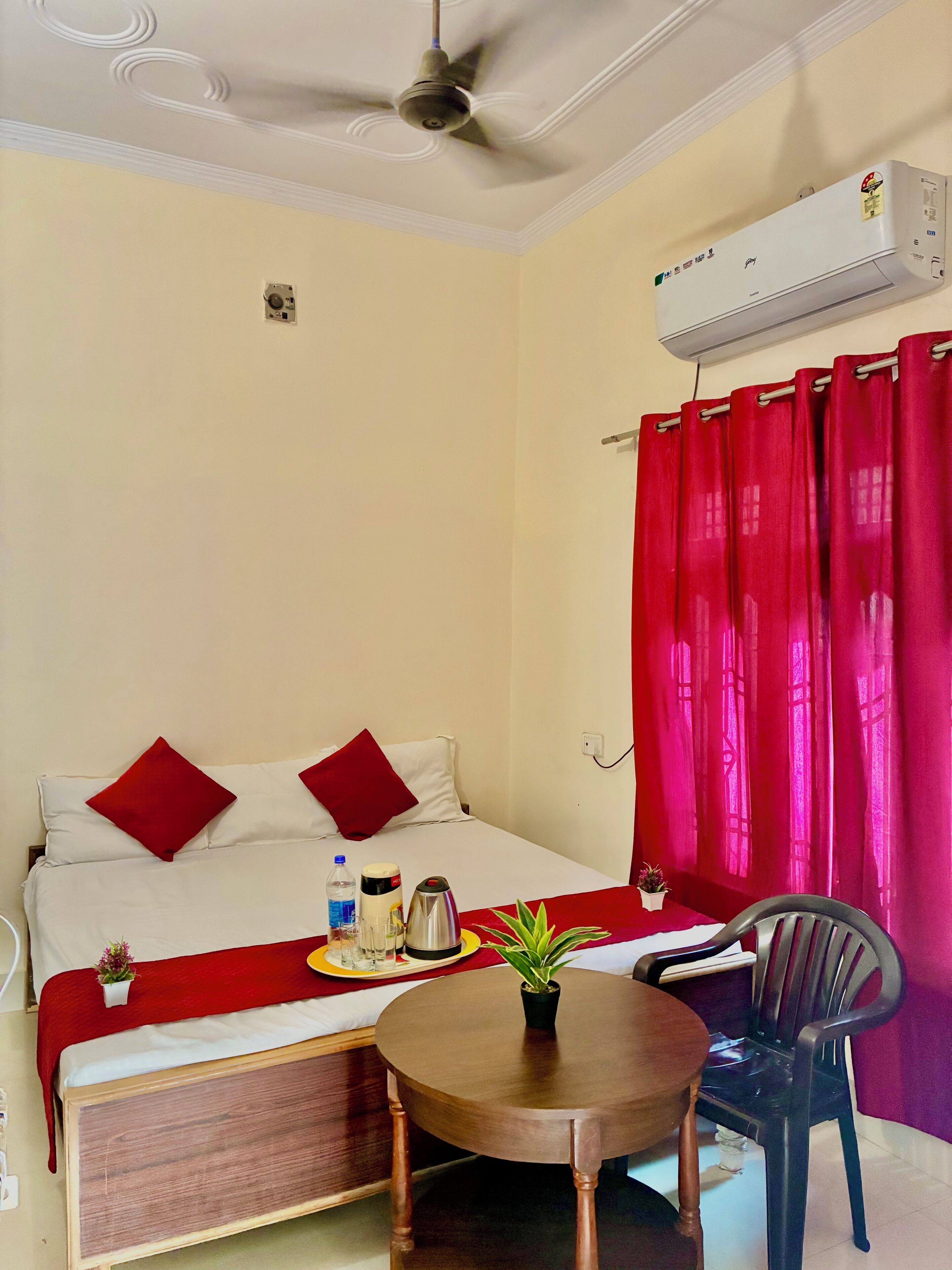 A hotel room with a bed, a ceiling fan, an air conditioning unit, a red curtain, a small table with a plant, and a chair.