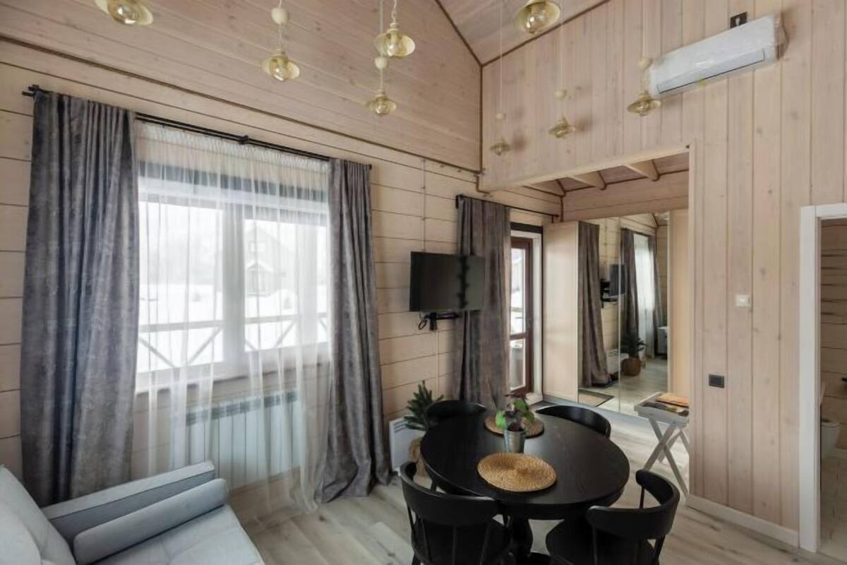 Een knus vertrek met een houten plafond, een ronde eettafel met stoelen, een bank en een aan de muur bevestigde televisie.