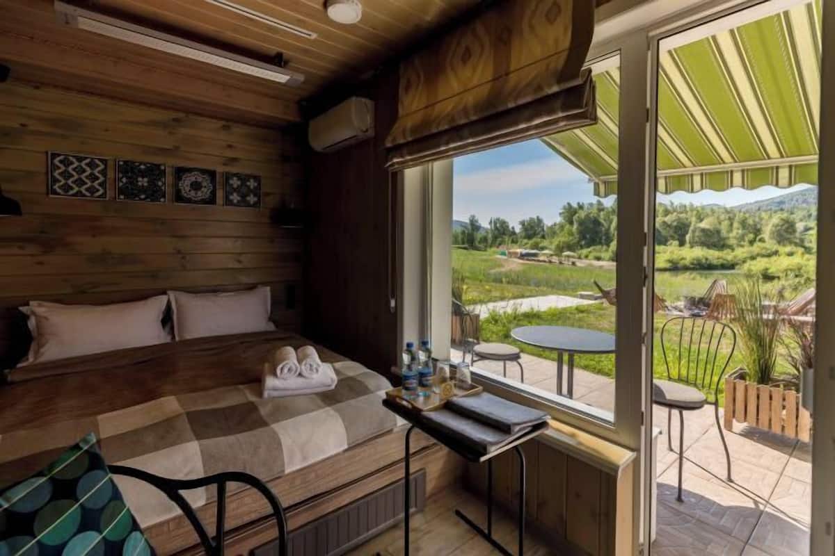 Een houten kamer met een bed, een tafel met handdoeken en uitzicht op een tuin en bergen.