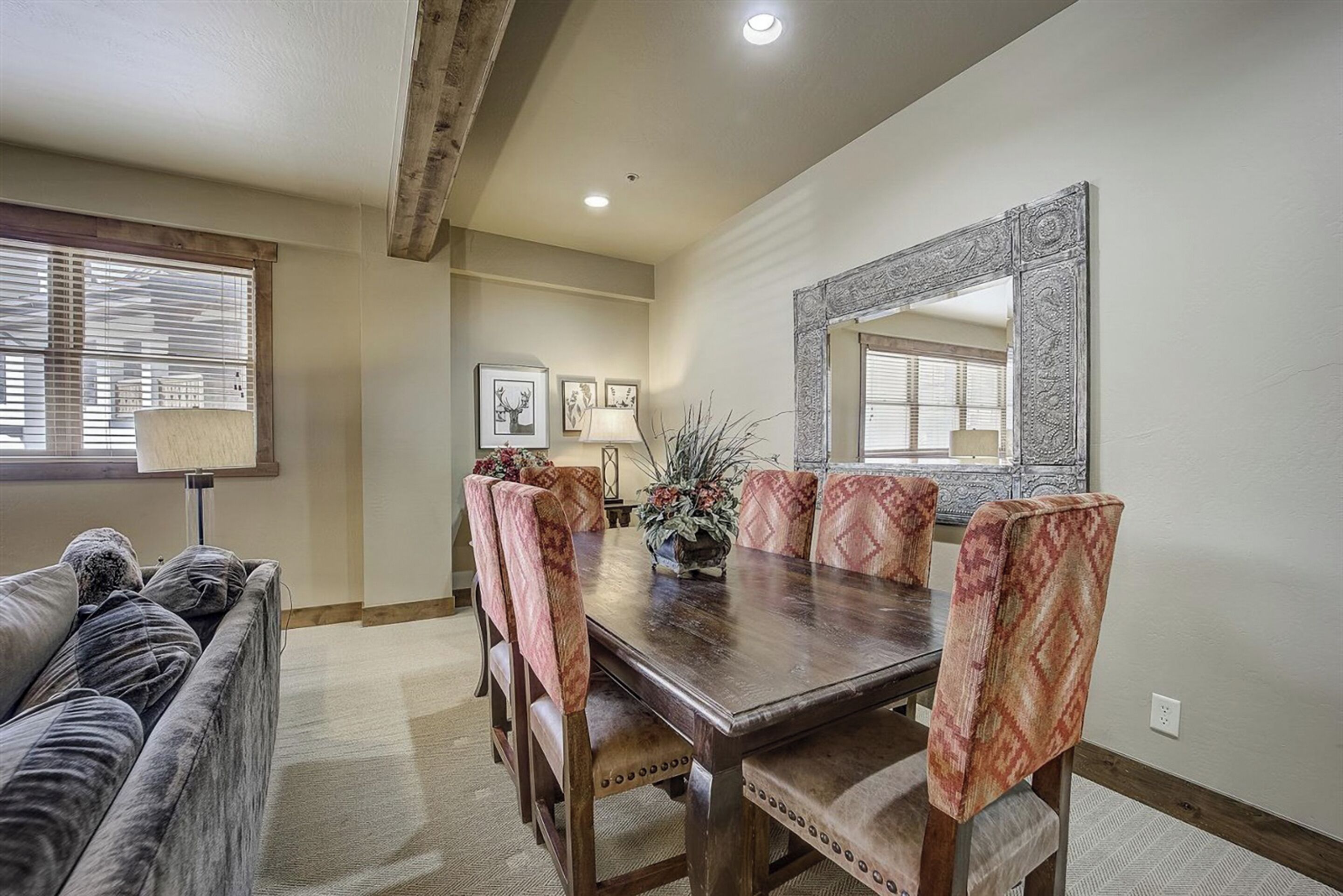 A dining area with a wooden table, patterned chairs, a sofa, and a large mirror on the wall.