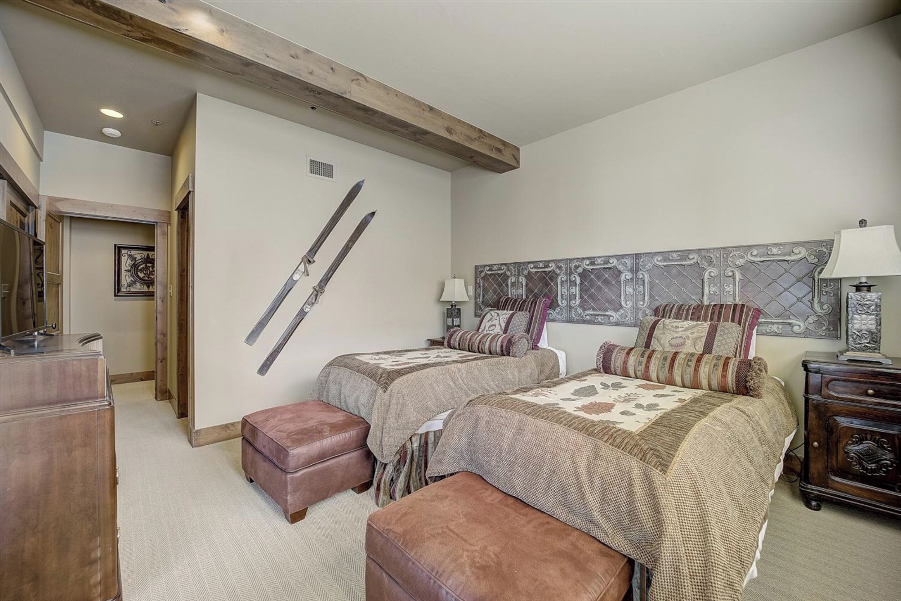 Een slaapkamer met twee bedden, een houten balk in het plafond, een ski-houder aan de muur en een nachtkastje met een lamp.
