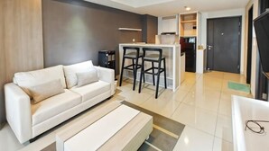 A modern living room with a sofa, a coffee table, and a kitchen area with a bar and stools. - Newly 1BR Apt Cikarang with Pool Gym & Wi-Fi (Bekasi Regency)
