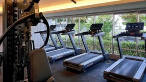A gym with treadmills, a weight machine, and a large window with a view of greenery. - Newly 1BR Apt Cikarang with Pool Gym & Wi-Fi (Bekasi Regency)