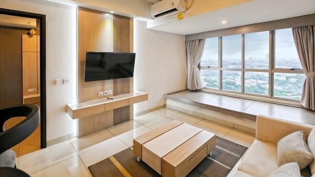 A modern living room with a flat-screen TV, a sofa, a coffee table, and a large window offering a city view. - Newly 1BR Apt Cikarang with Pool Gym & Wi-Fi (Bekasi Regency)