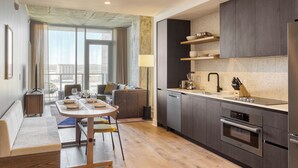 Een moderne keuken met donkere kasten, een inbouwoven en een spoelbak. Er is een eethoek met een tafel en stoelen, en een groot raam met uitzicht op de stad.