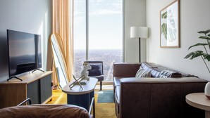 A modern living room with a large TV, a sofa, a chair, and a view of the city through large windows.
