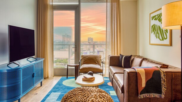 A modern hotel room with a large window, a sofa, a television, and a small table.
