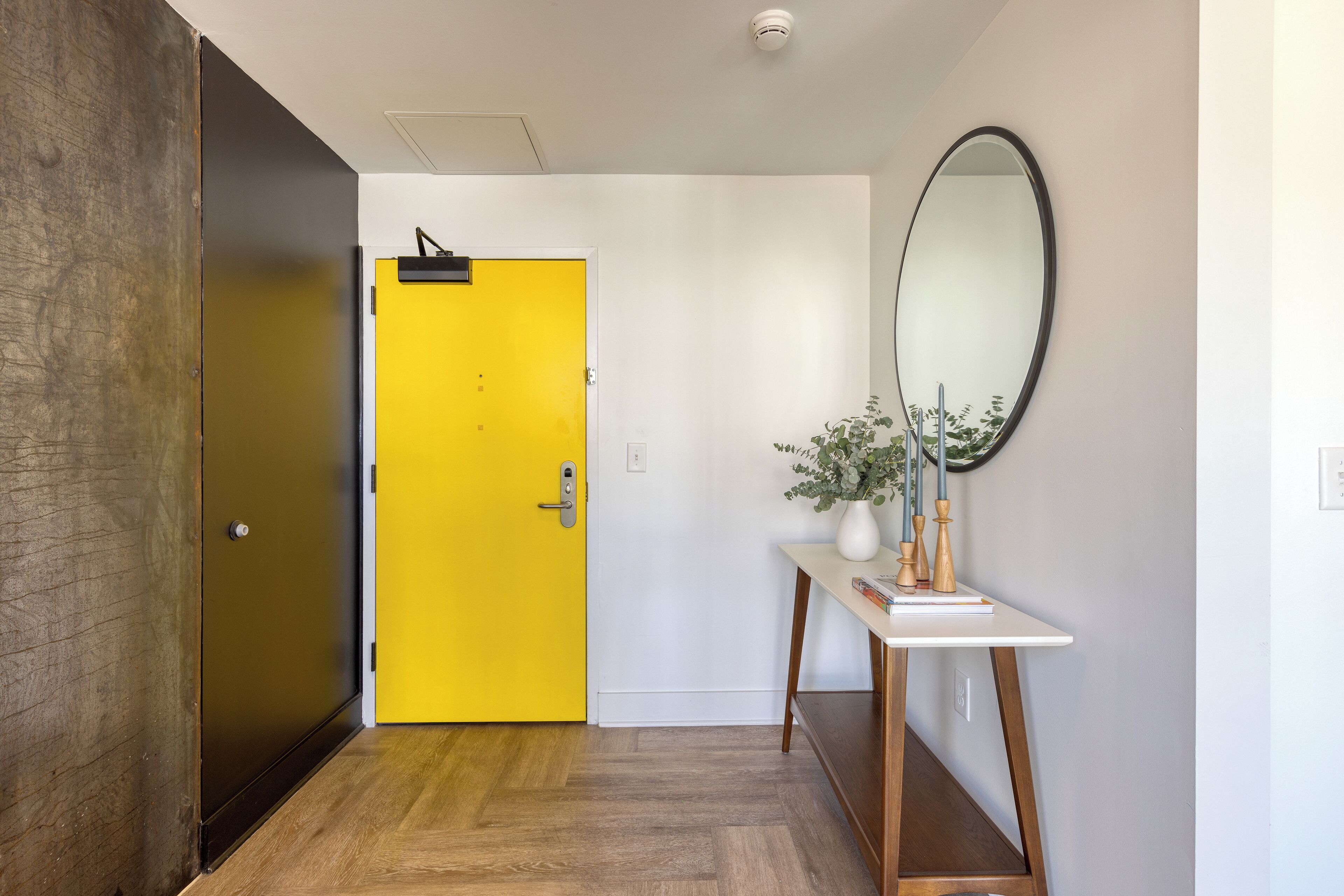 Un pasillo con una puerta amarilla, suelo de madera, pared blanca y un espejo.