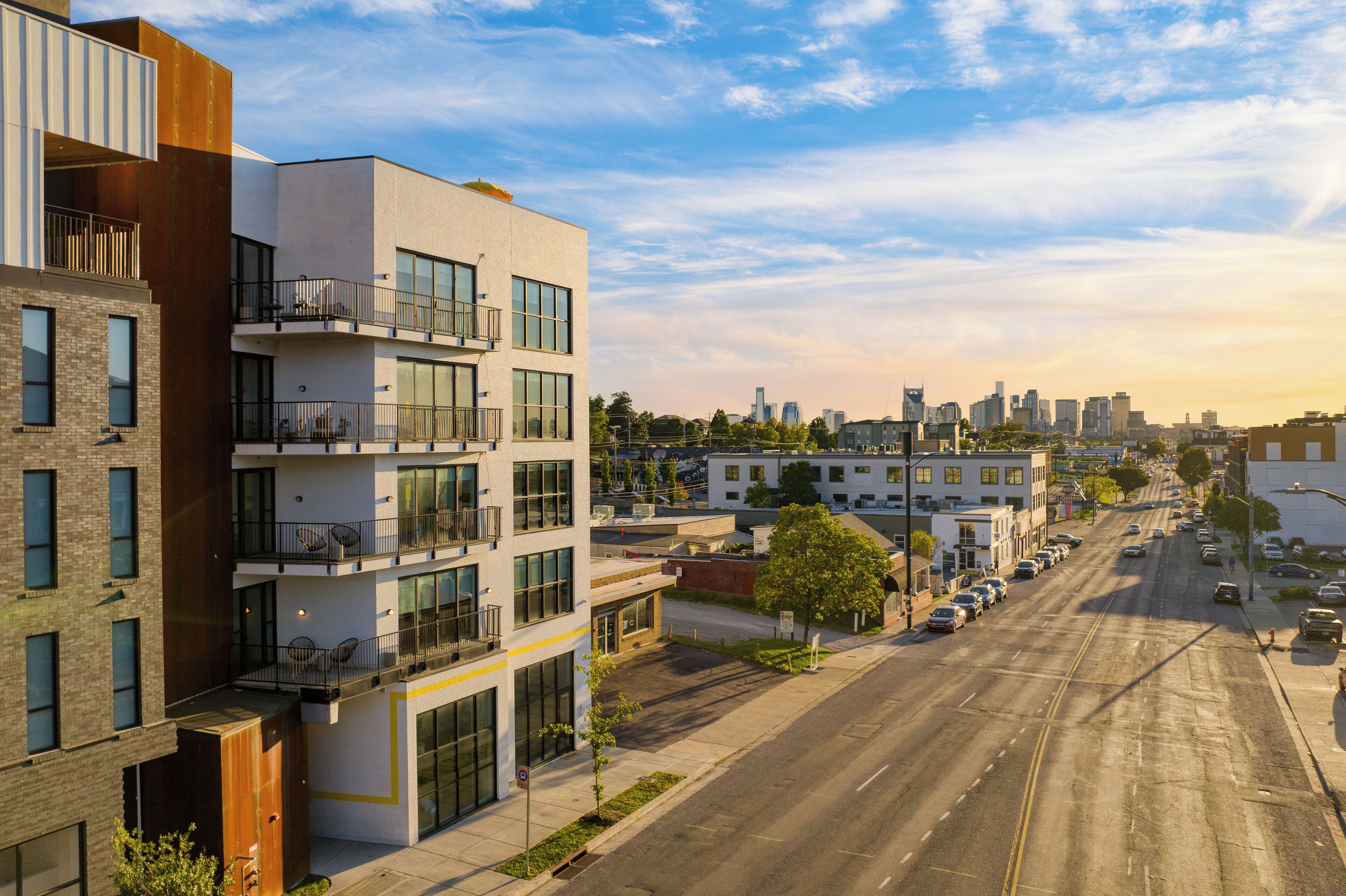 A modern multi-story building with balconies, a wide street, and a city skyline in the background.
