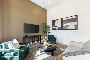 Ein modernes Wohnzimmer mit einem Flachbildfernseher, grünen Sesseln, einem runden Couchtisch mit einer Vase und Büchern sowie abstrakter Wandkunst.