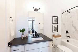 Ein modernes Badezimmer mit einem schwarzen Waschtisch, weißen Schränken, einer weißen Toilette und einer Badewanne. Über dem Waschtisch hängt ein Spiegel, daneben ein Topf mit einer Pflanze und an der Wand hängen zwei gerahmte Bilder.