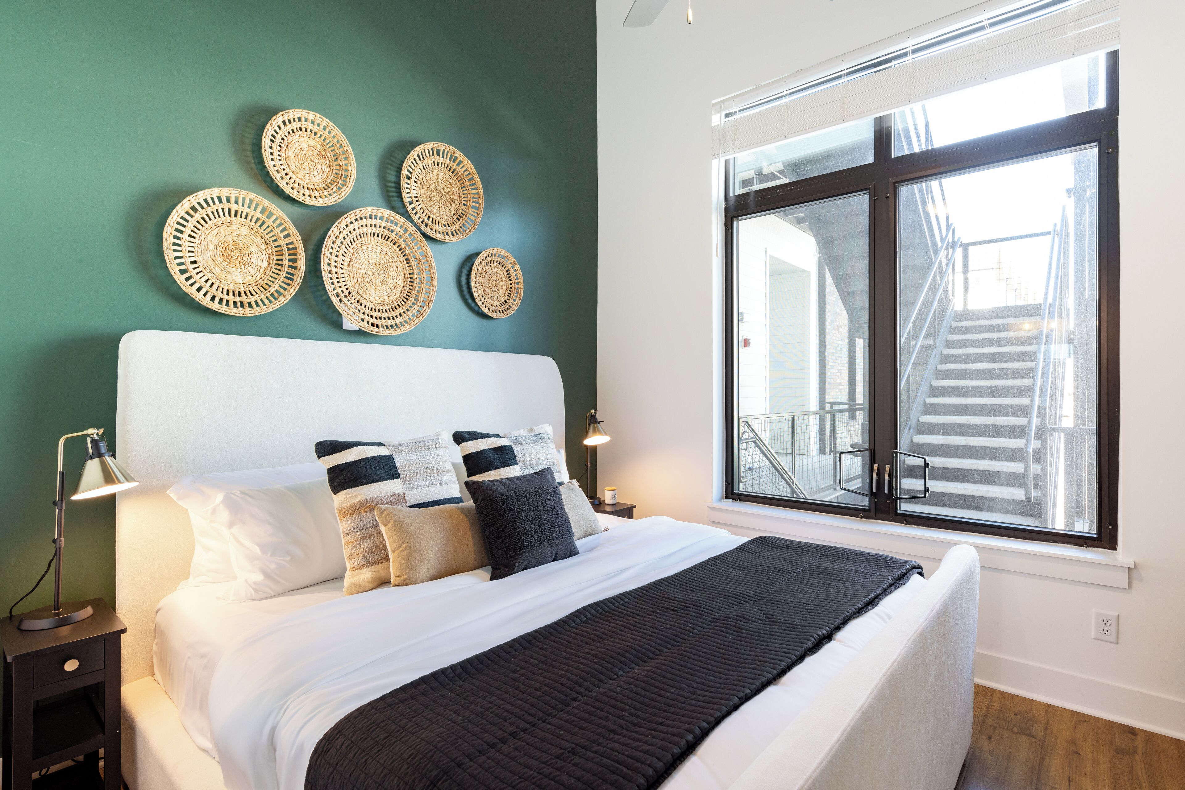 Un lit bien fait, avec une tête de lit blanche, un plaid noir et plusieurs coussins. Au-dessus du lit, on peut voir des décorations murales en fibres tressées, et une grande fenêtre donne sur un escalier.