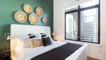 Un lit bien fait, avec une tête de lit blanche, un plaid noir et plusieurs coussins. Au-dessus du lit, on peut voir des décorations murales en fibres tressées, et une grande fenêtre donne sur un escalier.