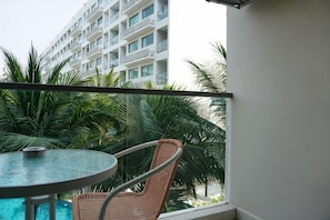 Varanda com mesa redonda e cadeira de vime, vista para uma piscina e palmeiras, com um edifício de hotel ao fundo.