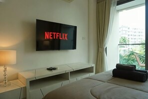 Habitación de hotel con una televisión en la pared mostrando 'NETFLIX', una cama, una mesita de noche con una lámpara y una ventana con vistas a edificios y vegetación.