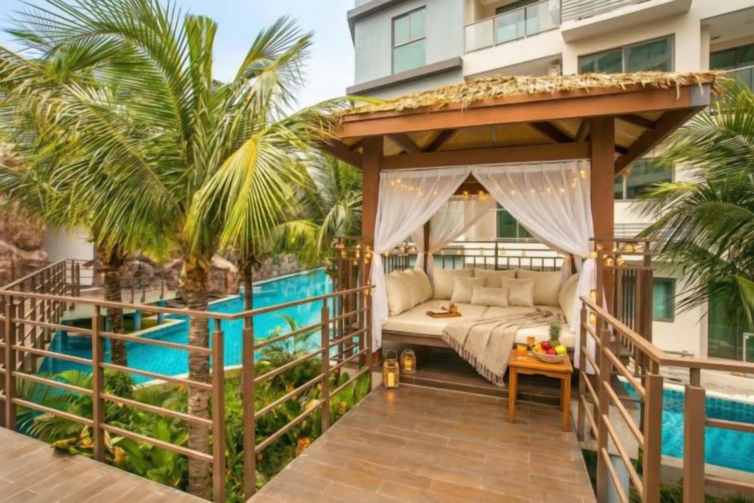 A private balcony with a wooden deck, a cushioned seating area, a small table, and a view of a swimming pool.