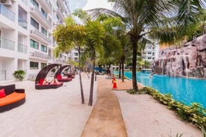 Un espace piscine d’hôtel avec un chemin en béton, des palmiers et une formation rocheuse.
