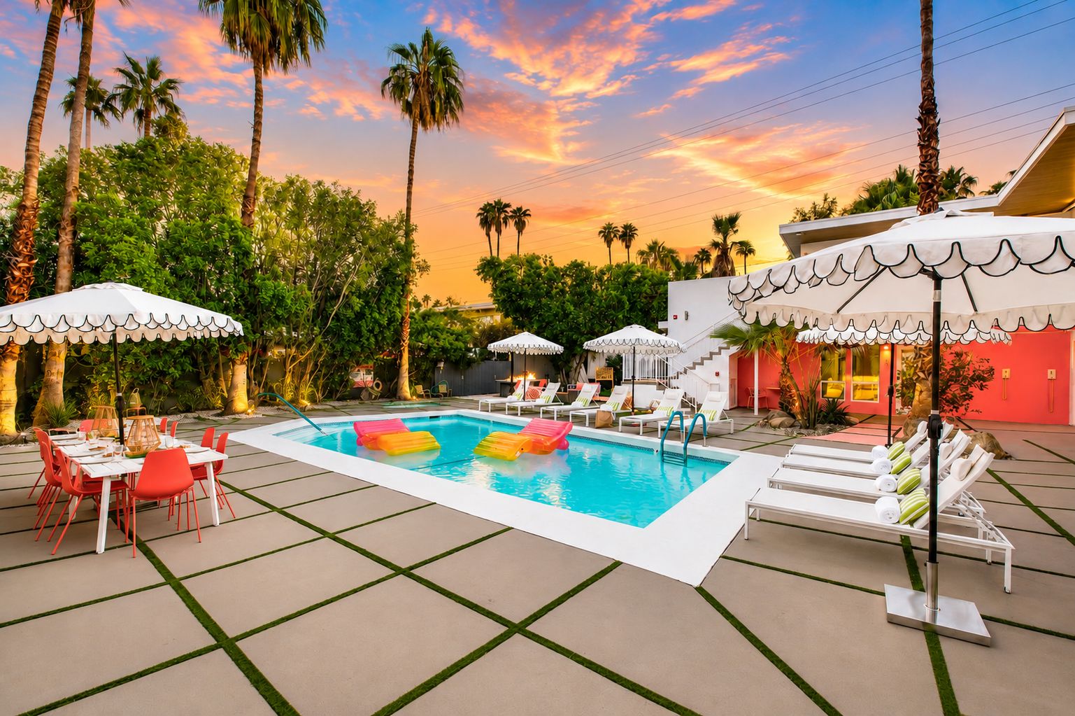 A swimming pool with lounge chairs and umbrellas, surrounded by palm trees and a sunset sky.