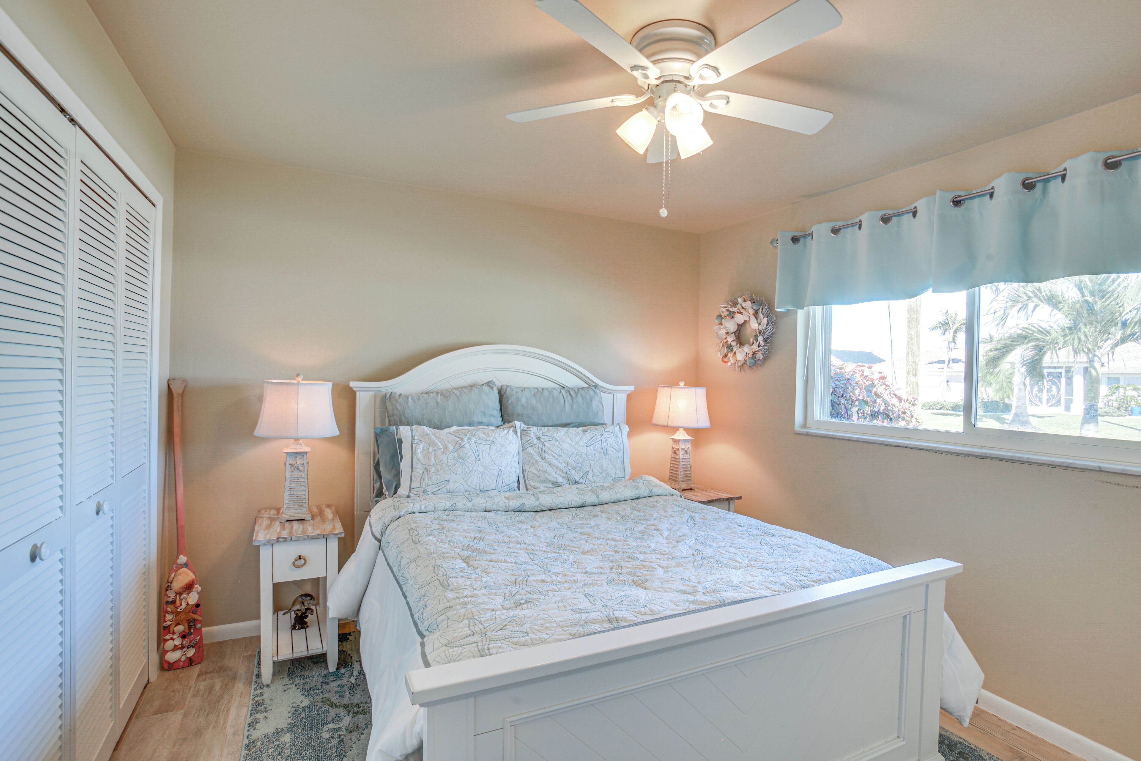 A bedroom with a bed, two bedside tables, a ceiling fan, a window with a curtain, and a wall-mounted decoration.