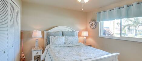 A bedroom with a bed, two bedside tables, a ceiling fan, a window with a curtain, and a wall-mounted decoration.