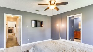 A bedroom with a bed, a ceiling fan, a TV, and a hallway leading to a bathroom. - Fish, Golf, Explore: Home 3 Mi to Prien Lake Park! (Lake Charles)