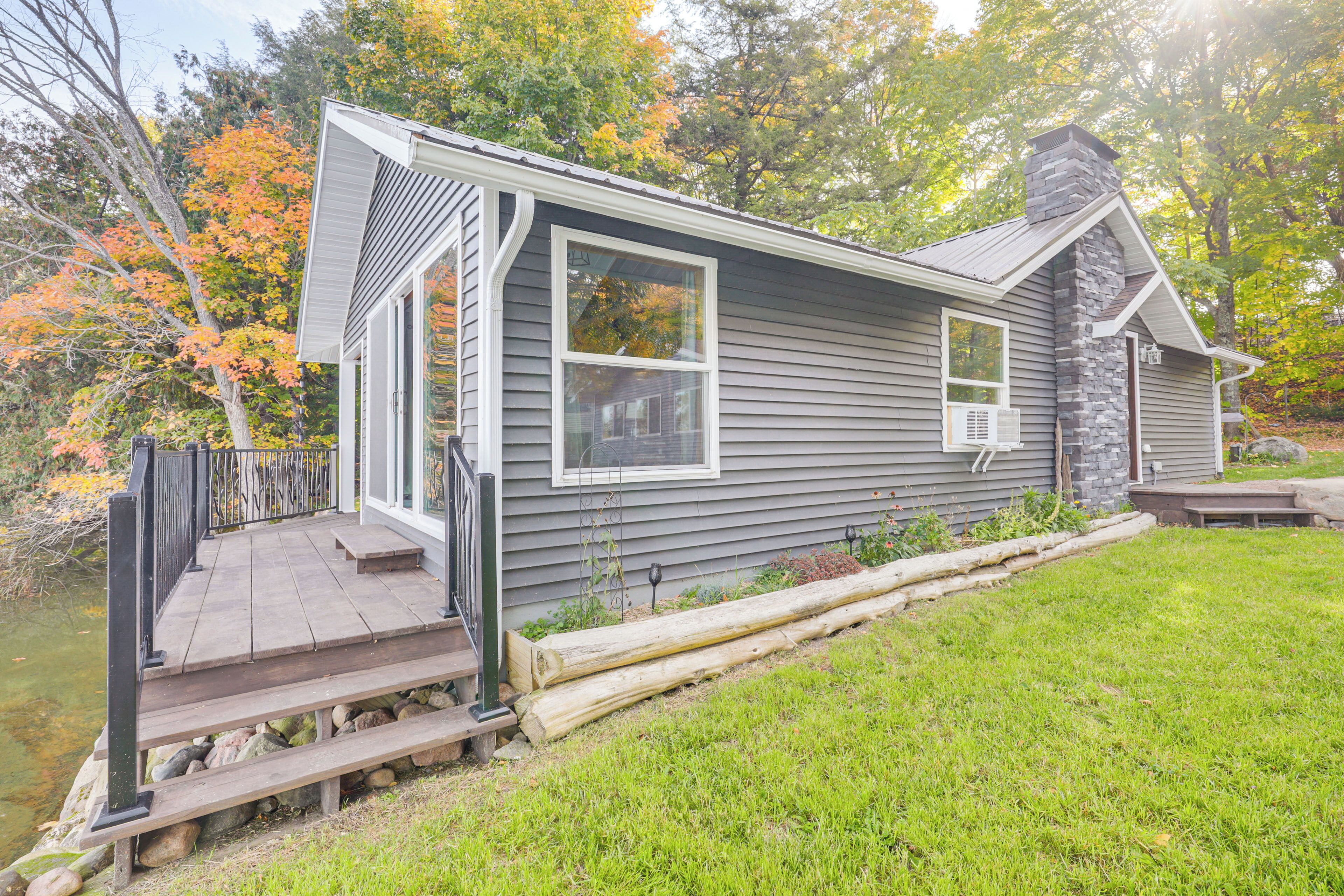 Une maison avec une cheminée en pierre, une terrasse en bois et un étang en arrière-plan.