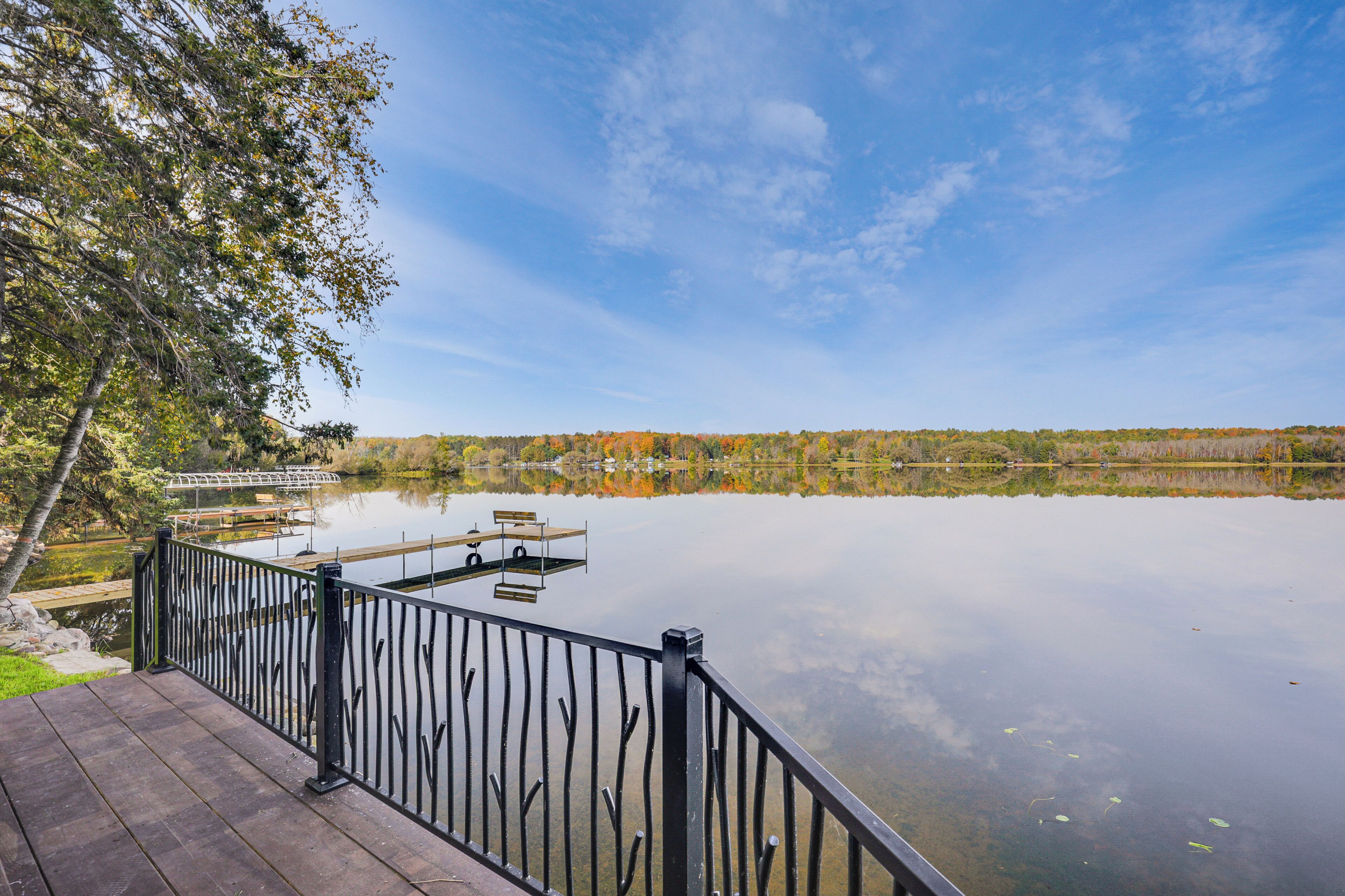 A wooden deck with a black railing overlooking a calm lake, surrounded by trees with autumn foliage.