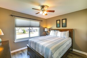 Un dormitorio con cama, ventilador de techo, ventana con persianas y dos cuadros en la pared.