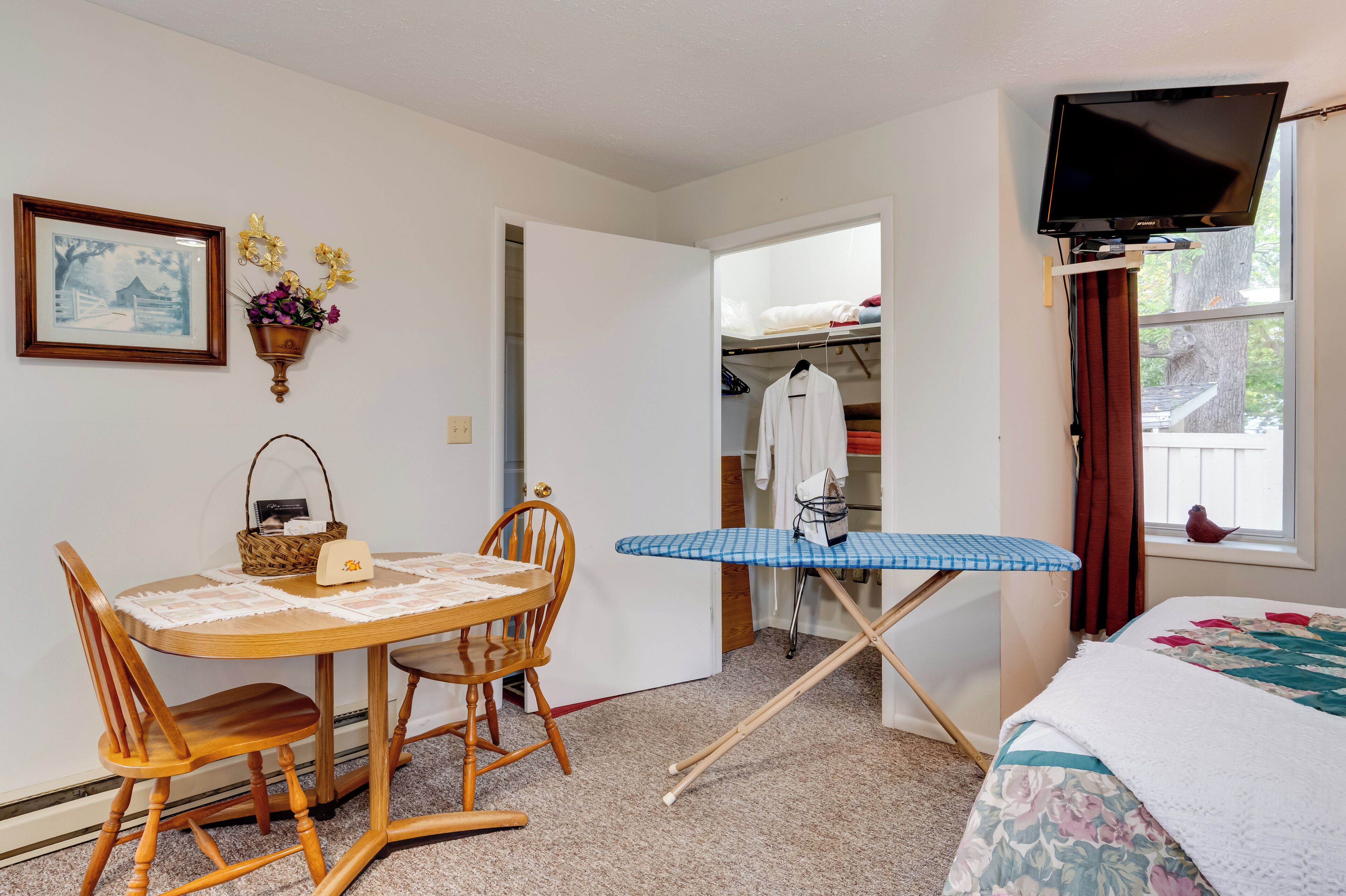 A room with a wooden dining table, chairs, a blue ironing board, a bed with a floral comforter, a TV on a stand, a closet with hanging clothes, and a framed picture on the wall.