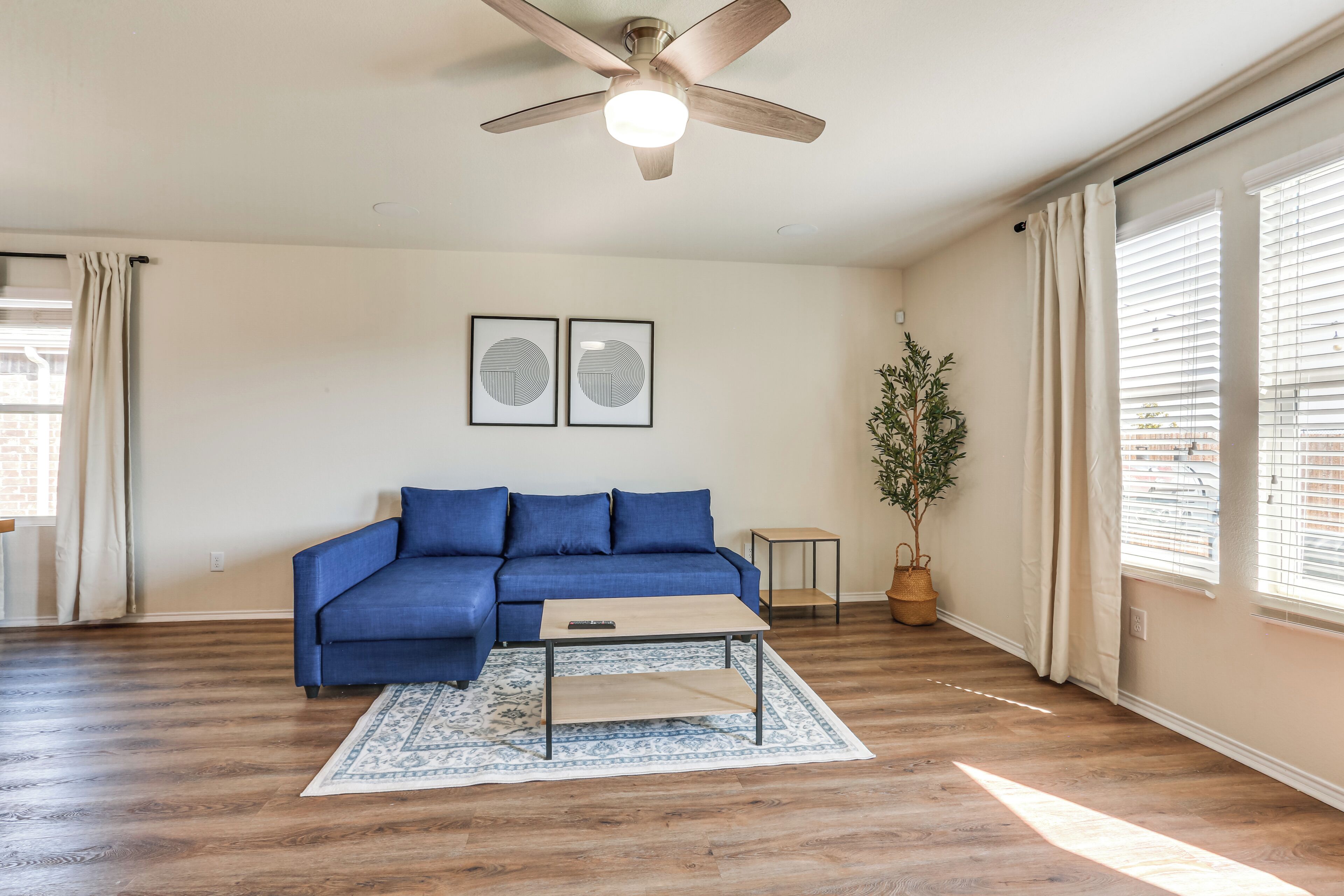 Un salon avec un canapé bleu, une table basse en bois, un tapis à motifs, un ventilateur de plafond et une plante en pot.
