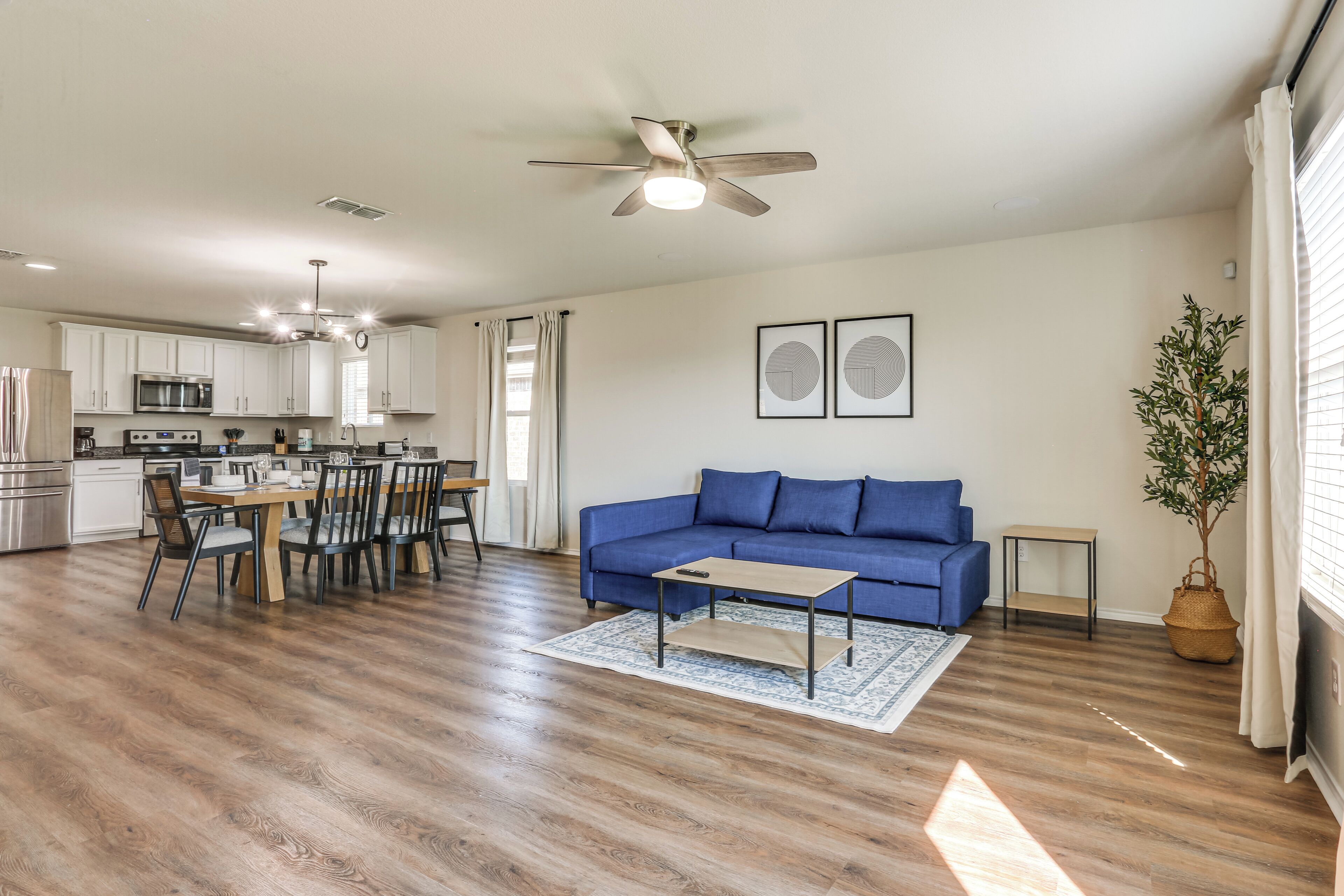 A modern living room with a blue sofa, wooden dining table, and chairs. A ceiling fan is visible, and there is a small side table with a plant.