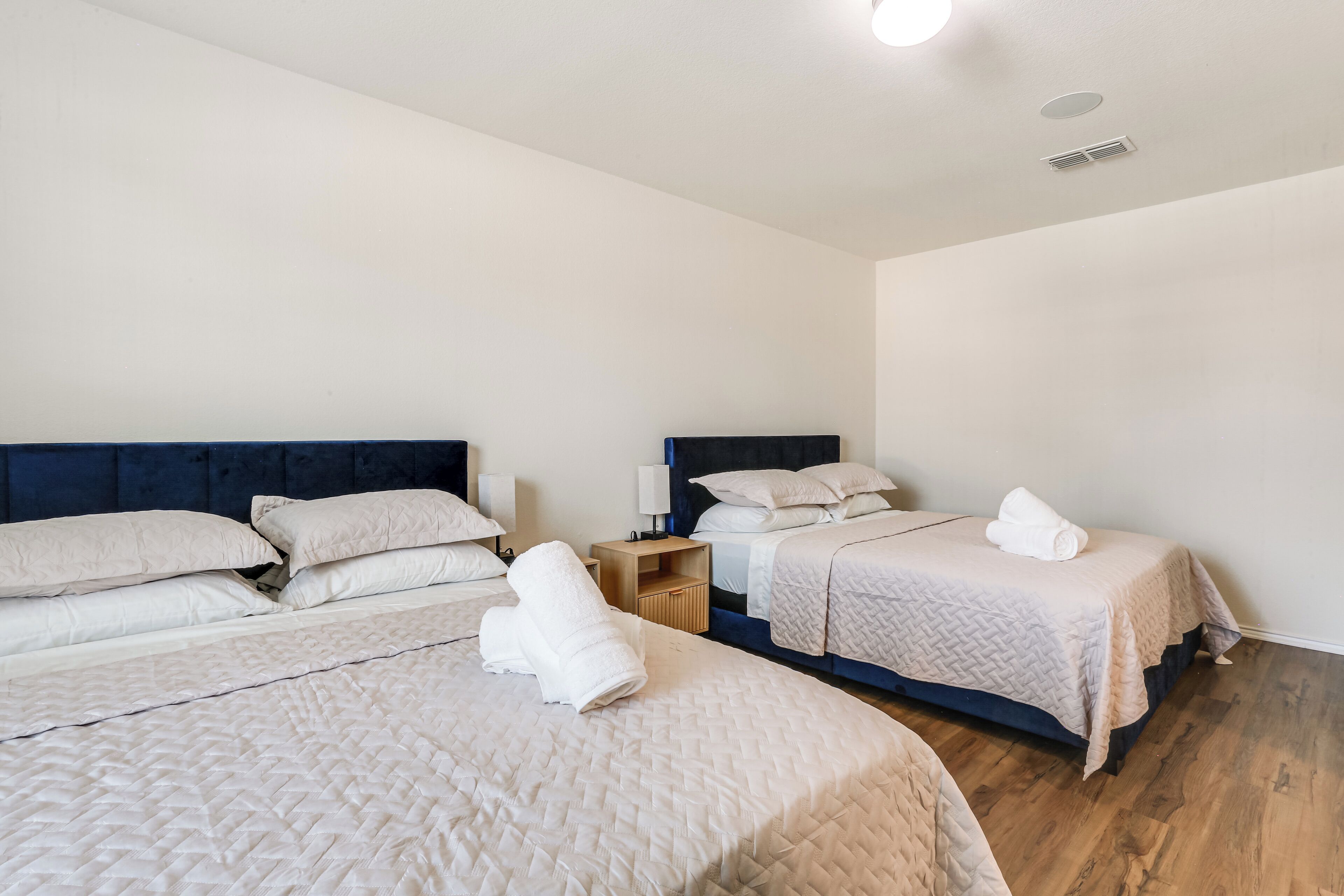 A hotel room with three beds, each with a headboard and pillows. There is a nightstand with a lamp and a small table.