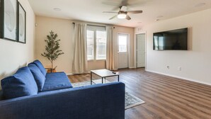 A living room with a blue sofa, a small table, a ceiling fan, a TV mounted on the wall, and a door leading to another room. - Modern Home w/ Private Yard & Patio in Little Elm! (Little Elm)