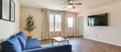 A living room with a blue sofa, a small table, a ceiling fan, a TV mounted on the wall, and a door leading to another room.